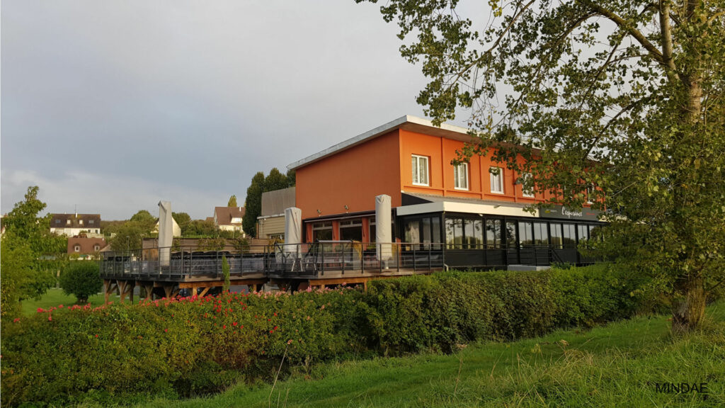 Cette photographie montre l'extérieur d'un bâtiment commercial à deux niveaux, probablement un restaurant nommé « L'espérance » comme le suggère l'enseigne noire sur la droite. L'étage supérieur arbore une façade orange vif percée de plusieurs fenêtres blanches alignées sous un toit plat. Au rez-de-chaussée, une grande structure moderne en verre et métal noir fait office de véranda ou de terrasse couverte panoramique. L'ensemble de l'établissement est implanté derrière une épaisse haie végétale verdoyante parsemée de petites fleurs rouges et roses à gauche, tandis que le premier plan est occupé par une pelouse et un grand arbre feuillu encadre le côté droit de la scène, tandis qu'un ciel couvert et nuageux s'étend à l'arrière-plan.